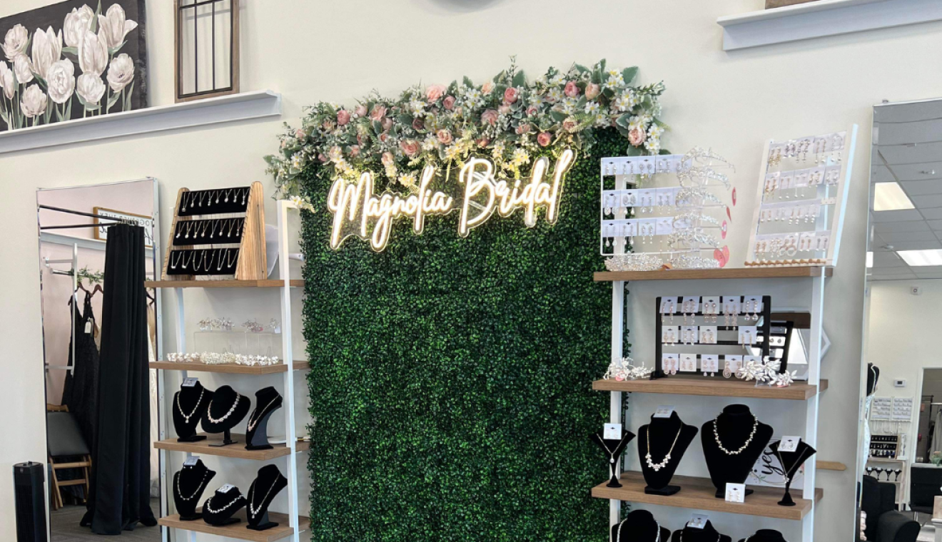 A photo of the interior of Magnolia Bridal, a bridal shop located near Grand Rapids. This image features a false greenery wall with an LED sign spelling out 'Magnolia Bridal' in cursive. There are two jewelry stands on either side of the greenery walls.