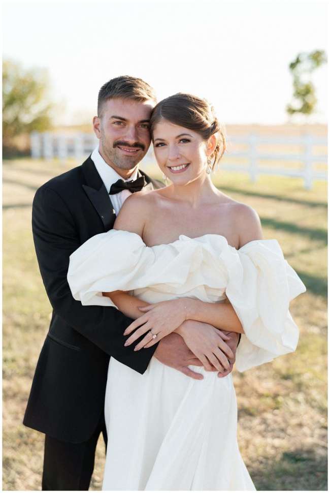 Groom hugs his bride as they both face the camera. The bride has a lush ivory dress with large sleeves and pearl drop earrings. 